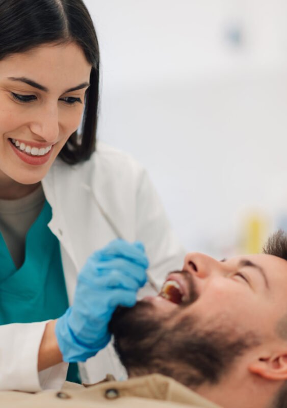 Smiling female dentist wearing gloves examines the teeth of a male patient reclining in a dental chair within a bright, modern dental clinic, ensuring optimal oral health and hygiene