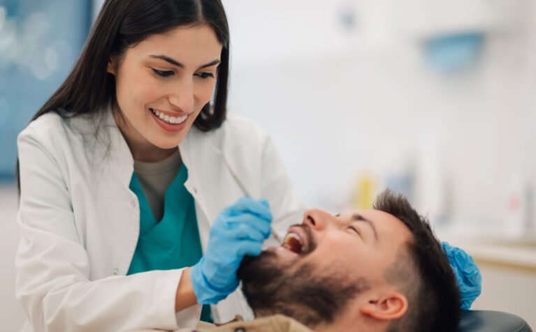 Smiling female dentist wearing gloves examines the teeth of a male patient reclining in a dental chair within a bright, modern dental clinic, ensuring optimal oral health and hygiene