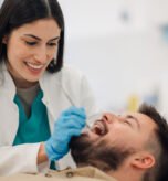 Smiling female dentist wearing gloves examines the teeth of a male patient reclining in a dental chair within a bright, modern dental clinic, ensuring optimal oral health and hygiene