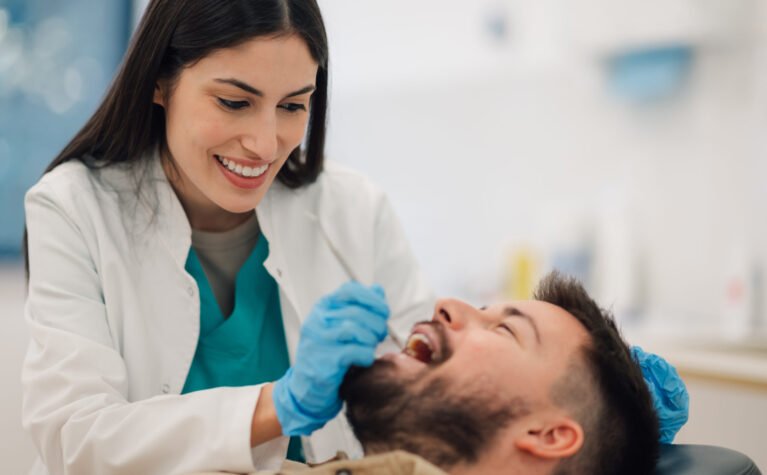 Smiling female dentist wearing gloves examines the teeth of a male patient reclining in a dental chair within a bright, modern dental clinic, ensuring optimal oral health and hygiene
