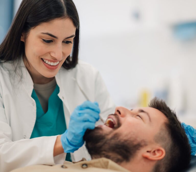 Smiling female dentist wearing gloves examines the teeth of a male patient reclining in a dental chair within a bright, modern dental clinic, ensuring optimal oral health and hygiene